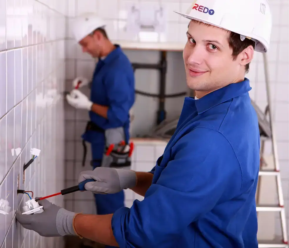 REDO technician in blue uniform and hardhat performing electrical and plumbing work on a wall